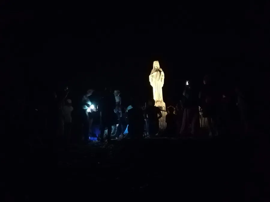 A floodlit statue of Our Lady on Apparition Hill surrounded by people in complete darkness