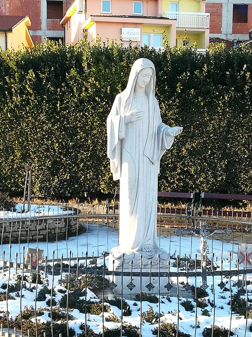 A white statue of Our Lady in a railed garden with snow lying on the ground and village houses behind